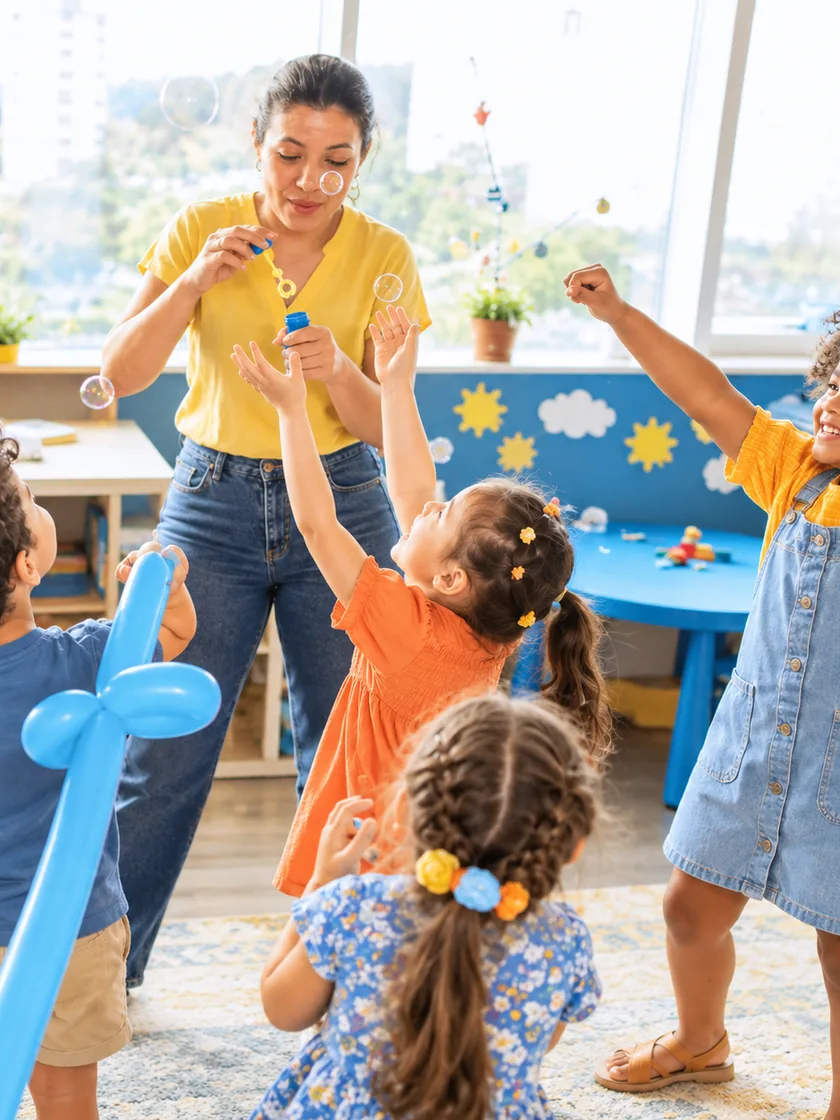 A teacher blowing bubbles for a group of children in a bright playroom, two of them reaching up for one
