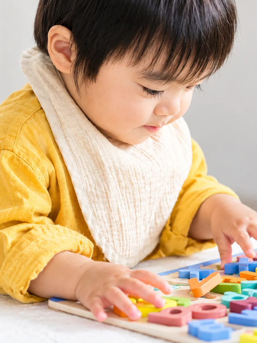 A toddler in a mustard shirt leaning over a wooden alphabet puzzle, fitting a letter into place