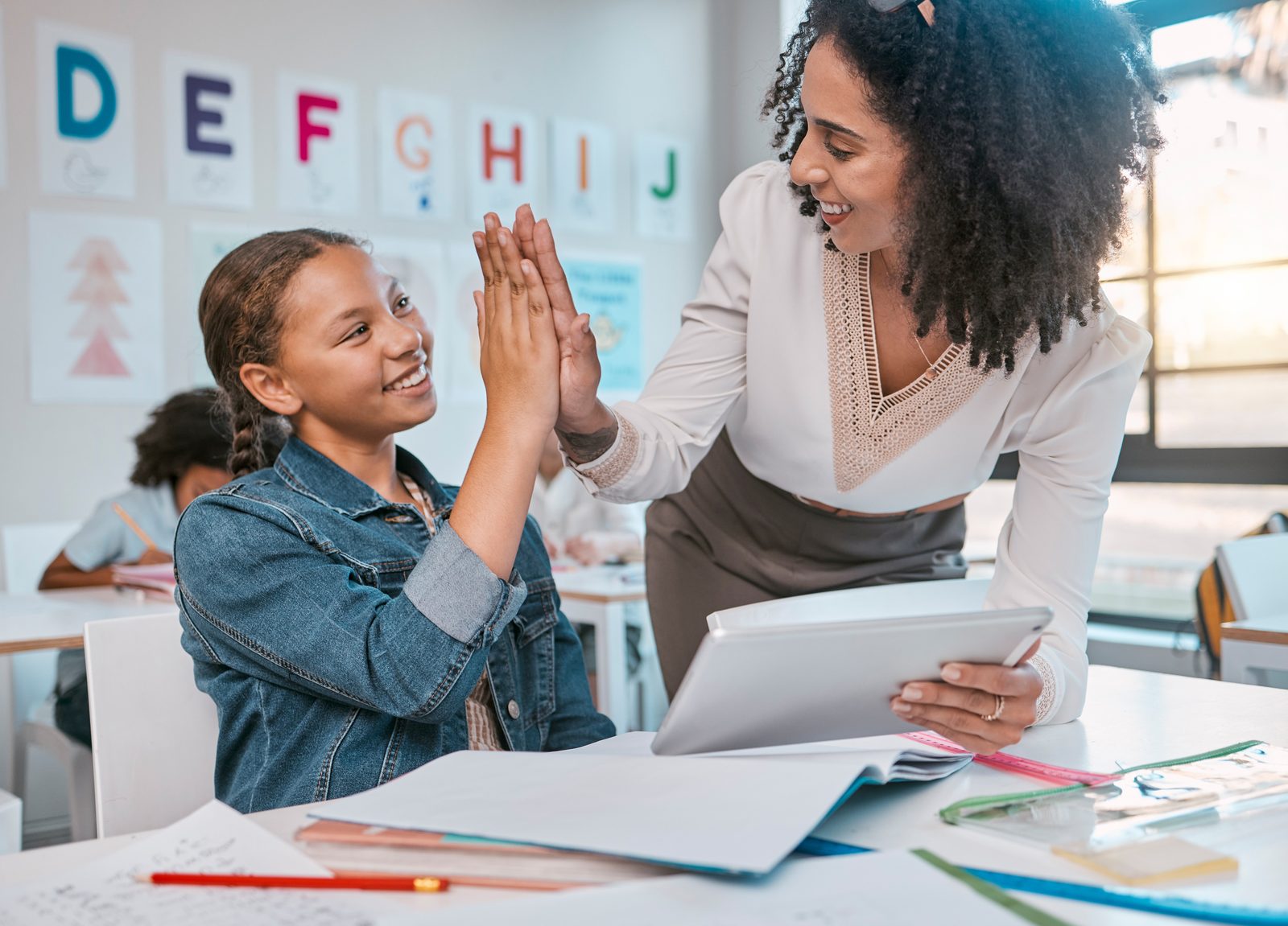 A teacher high-fiving a smiling girl at her desk in a bright classroom, a tablet in her other hand