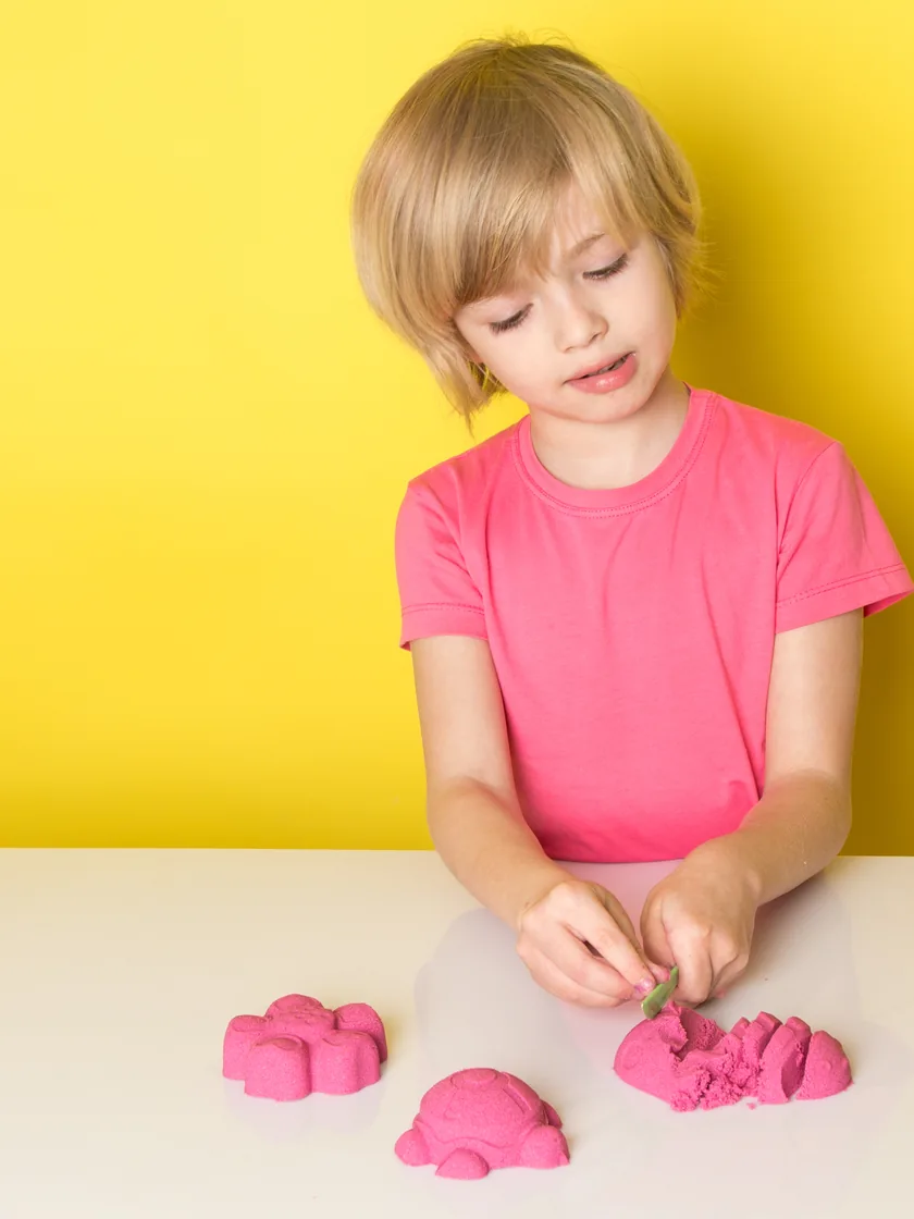 A child in a pink t-shirt pressing shapes out of pink kinetic sand at a table