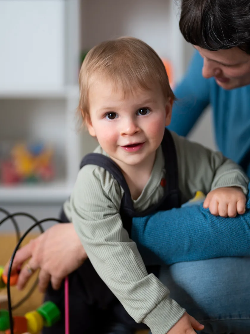A toddler in dungarees smiling at the camera while a parent sits beside them at a bead maze