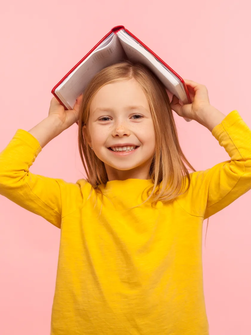 A smiling child holding an open book over their head like a roof
