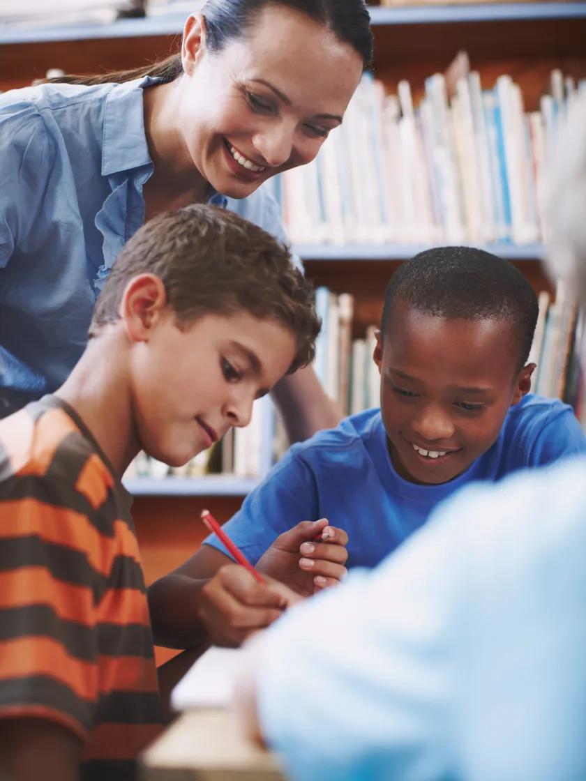 A teacher leaning over two children writing at a table in front of a wall of library shelves