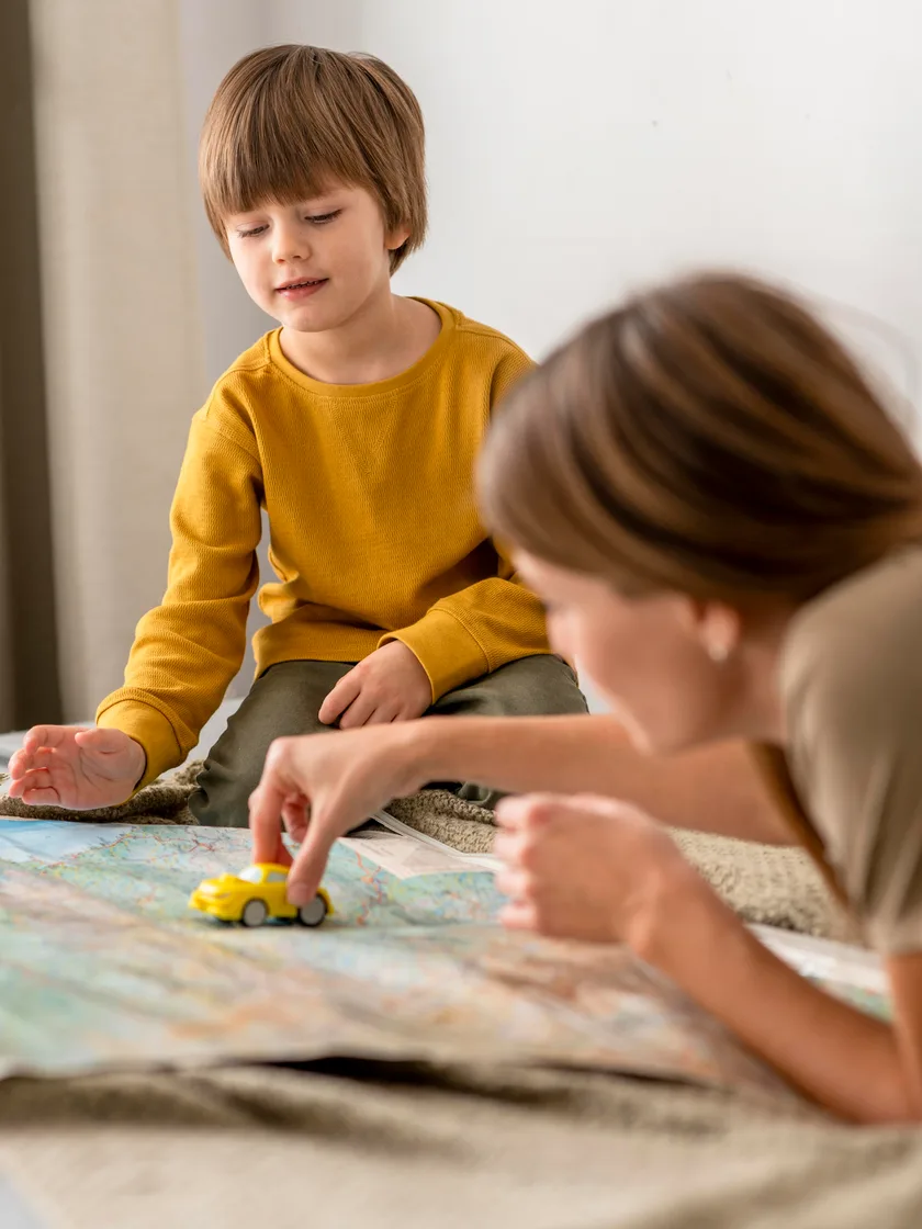 A boy in a mustard jumper watching a parent push a small yellow toy car across a map on the floor