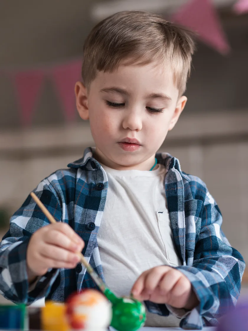 A young boy in a checked shirt concentrating on painting an egg with a fine brush