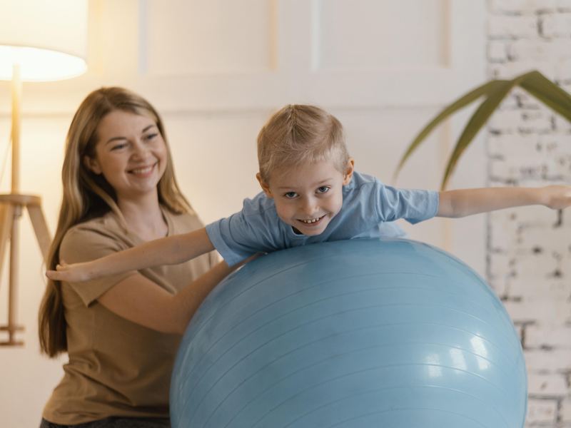 A boy stretching both arms wide over a large exercise ball while an adult steadies him