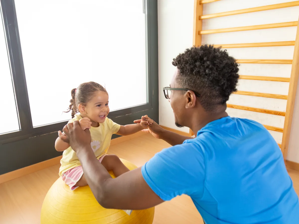A man in a blue T-shirt holding a laughing young girl's hands as she balances on a large yellow exercise ball in a bright therapy room