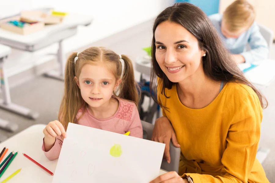 A girl holding up a drawing of a sun and hearts beside her smiling teacher at a classroom table