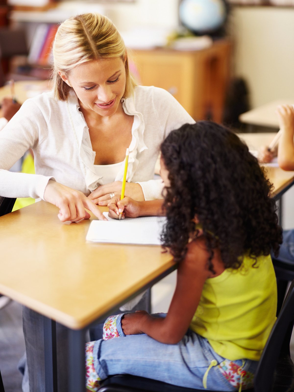 A smiling teacher pointing to a notebook while a girl with curly hair writes with a yellow pencil at a classroom desk